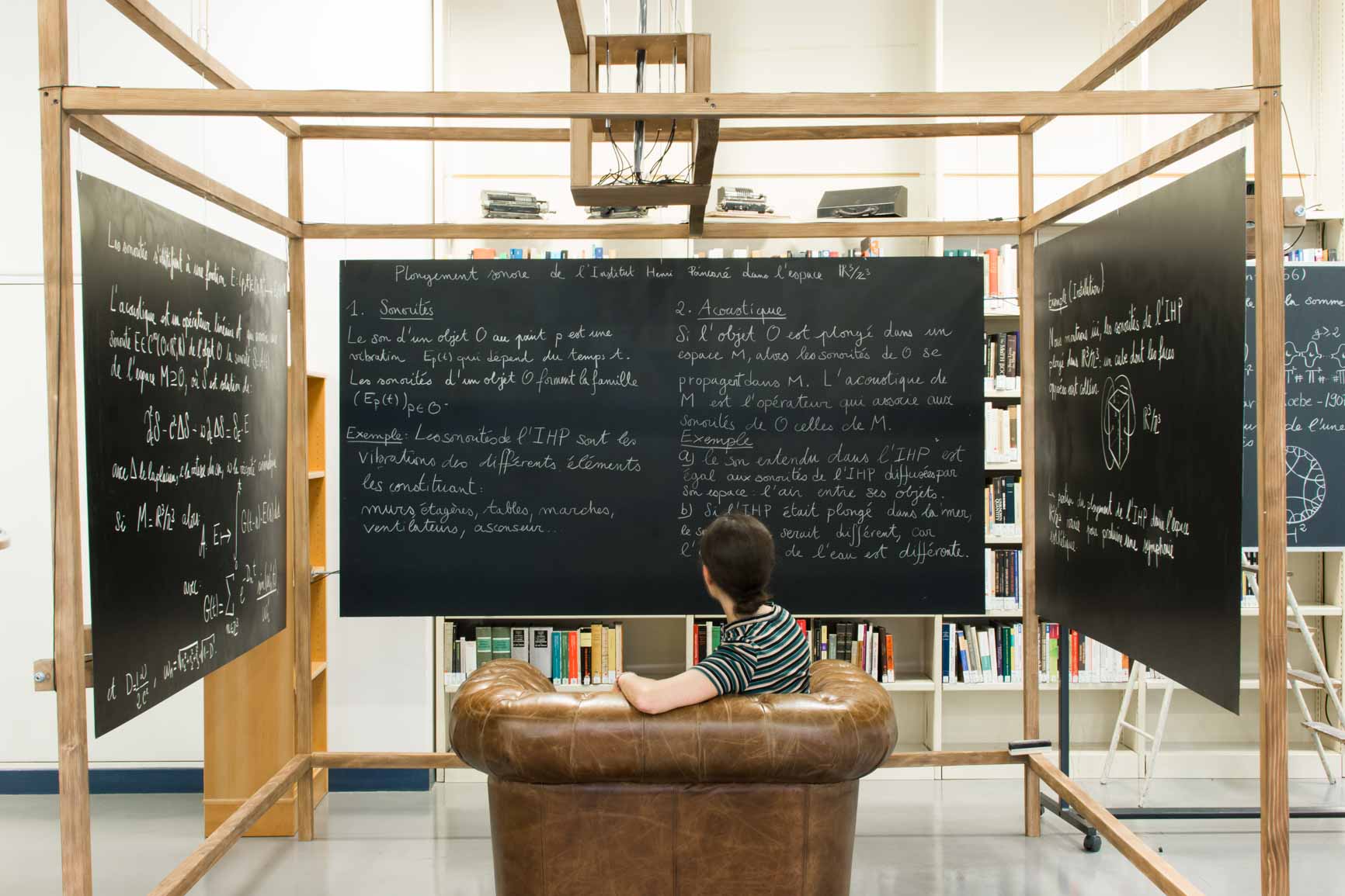 Deux tableaux à craie sont disposés face à face et le troisème se trouve entre les deux. Un canapé est disposé face au troisième et un visiteur est assis dessus en contemplant les tableaux